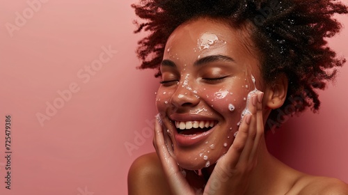 A youthful black girl with radiant skin cleanses her face using beauty product while grinning and gazing upwards, relishing the silky sensation of a moisturizing facial mask.