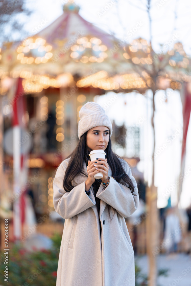 A thoughtful woman holding a takeaway coffee cup, gazing at a carousel with festive lights. 