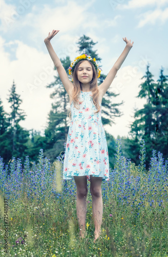 Full-length portrait of a beautiful girl with a flower wreath on her head, with closed eyes and raised hands enjoying the colorful spring field