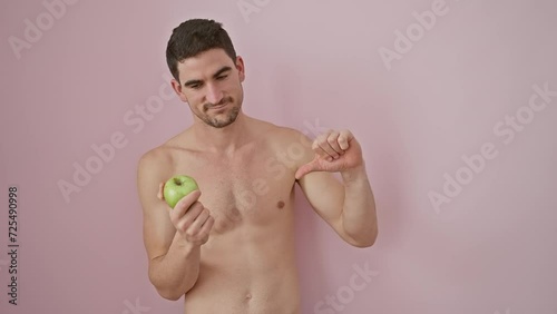 Shirtless young hispanic man, peeking in shock, coyly covering eyes with hand and an apple, peers in fear over a pink isolated backdrop photo