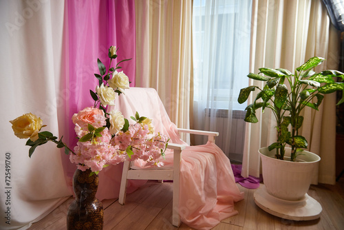Delicate bright pink interior of the room with armchair, a vase with roses, draped curtains and a window. Location and background in the photo studio
