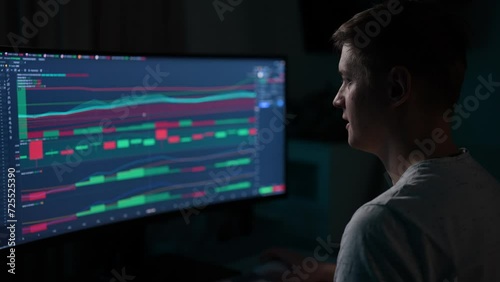 Focused financial male analyst using computer with display showing real-time stocks, exchange market charts. Remote working male stock trader making e-commerce investment sitting at desk in dark room. photo