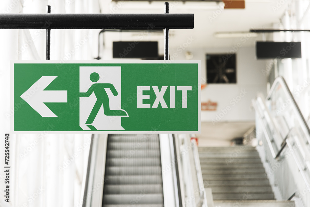 Emergency exit sign on the left in a railway station. Green safety sign with arrow in the foreground, blurred stairs on the background.