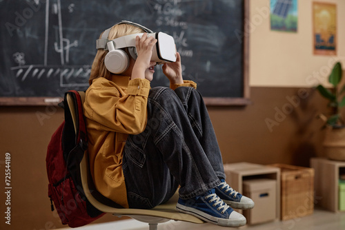 Full length side view of young blond boy wearing VR headset in class and enjoying high tech immersive learning experience