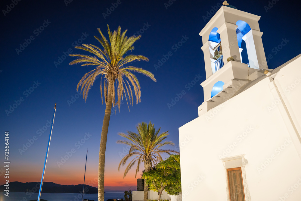 View of the illuminated  whitewashed Orthodox Church of Panagia  Gremniotissa in Ios Greece,  surrounded by palm trees at sunset