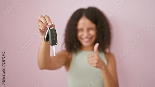 Cheerful young hispanic woman thrillingly holding up new car key, smiling confidently, her thumb up in joy, backed by a fun pink isolated wall. photo