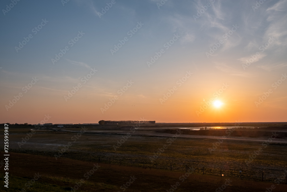 This image captures the tranquil beauty of a sunset over a rural landscape. The sun is seen nearing the horizon, casting a warm golden glow across the scene. A sparse expanse of flat land stretches