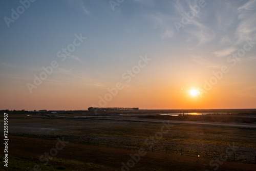 This image captures the tranquil beauty of a sunset over a rural landscape. The sun is seen nearing the horizon, casting a warm golden glow across the scene. A sparse expanse of flat land stretches