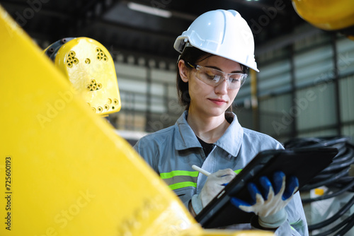 Caucasian female robotic engineer wearing helmet and safety glasses using tablet to check the operation of industrial robot arm in factory.