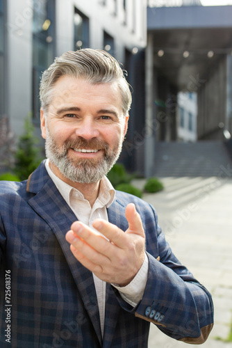 Cheerful experienced senior professional offering a handshake in a friendly gesture, standing confidently outside an office.