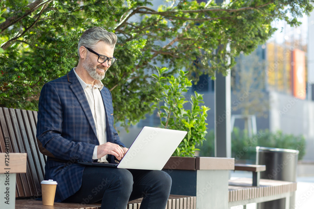 Mature businessman in a suit working on a laptop outside an office building, portraying professionalism and experience. Mature businessman in a suit working on a laptop outside an office building, portraying professionalism and experience.