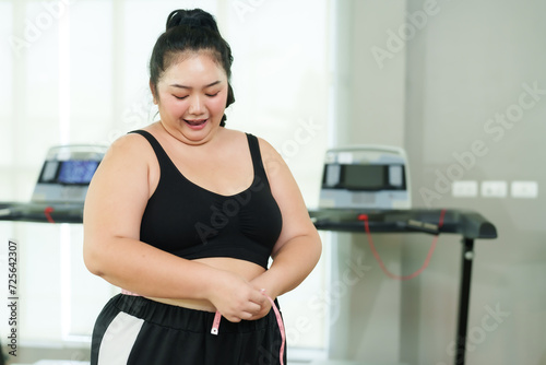 Overweight woman, beautiful Asian woman is using a measuring tape to measure waist size and expressing happiness at successful weight loss, standing in village gym. Lifestyle and exercise concepts