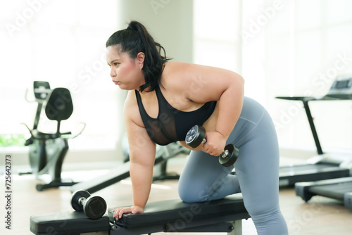 Overweight Asian woman exercise in gym, activewear engaged in a dumbbell workout on bench, concentrating on fitness regimen. Focused female performing dumbbell rows, dedication to workout routine