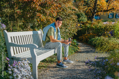 A Teenager Sits On A Bench In The Autumn Park Drinks Coffee From A Thermo Mug And Looks Into A Phone. Portrait Of Handsome Cheerful Guy Sitting On Bench Fresh Air Using Device Browsing Media Smm
