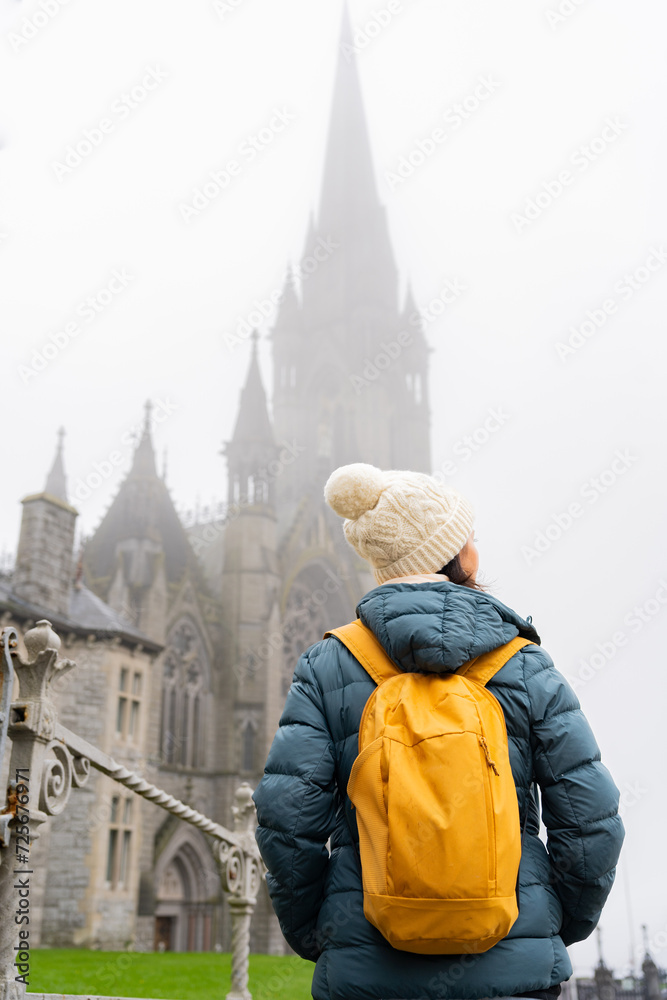 Unknown woman with knit hat and yellow backpack enjoying a beautiful and unique view of the cathedral of a small town in Ireland on a foggy day