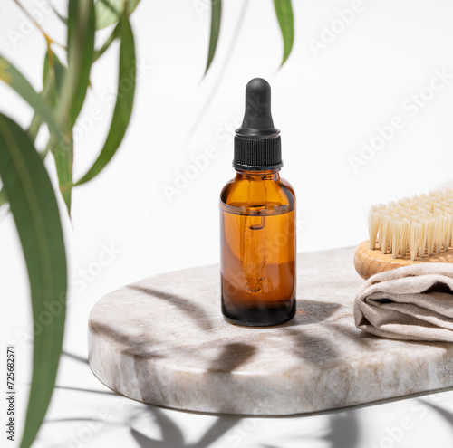 Nail oil or serum in a bottle on a marble board with a brush on a white background with green leaves and shadow.