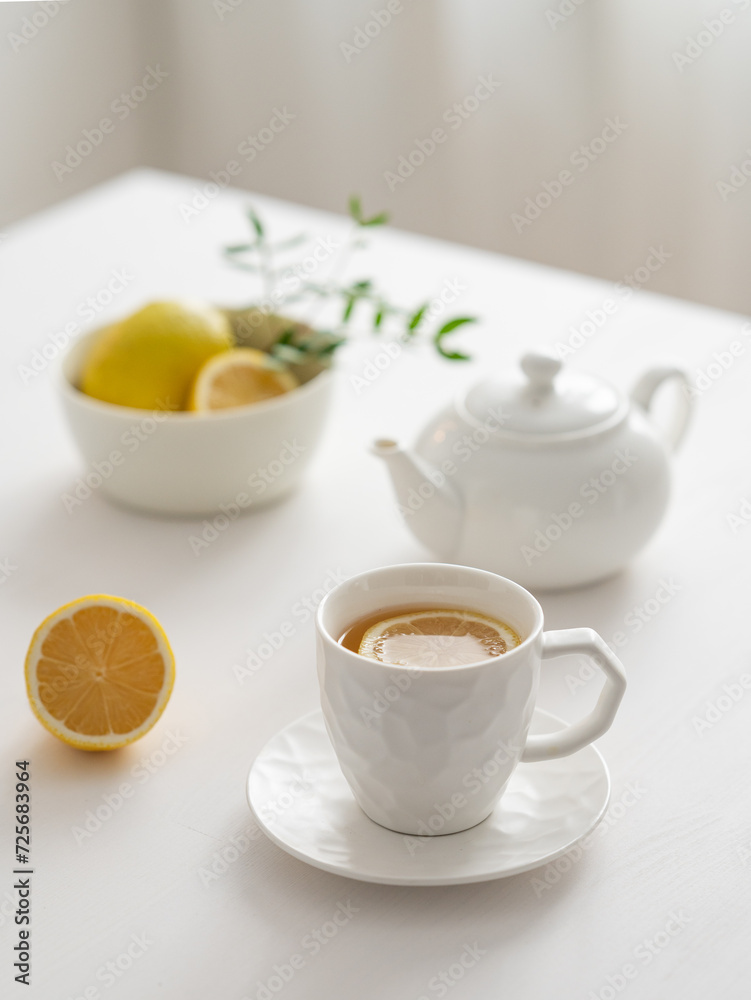 A cup of tea with lemon and a teapot on a white table against the background of a kitchen window. A cup of tea with lemon and a teapot on a white table against the background of a kitchen window.