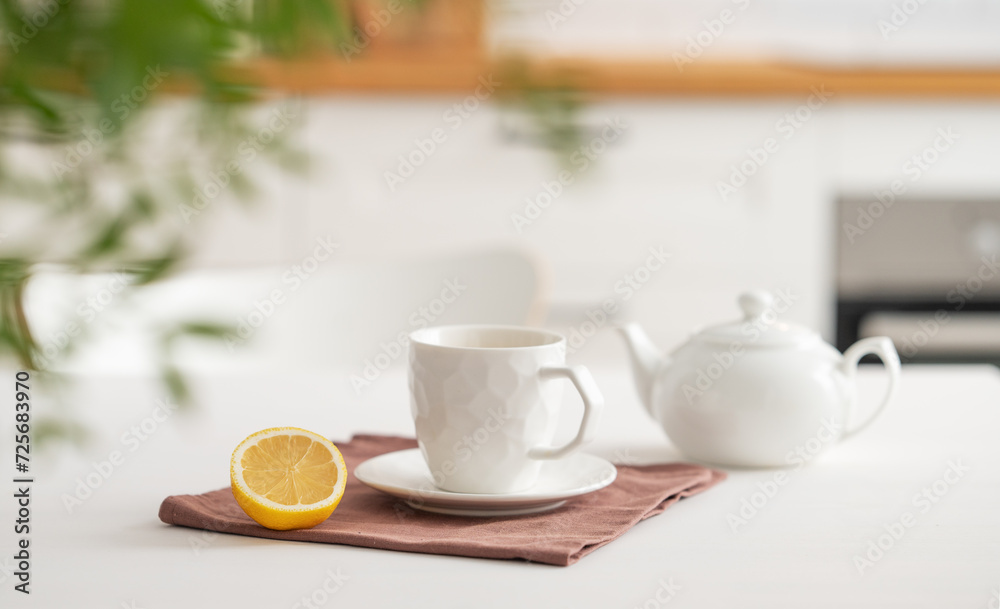 A cup of tea with lemon and a teapot on a white table with branch.