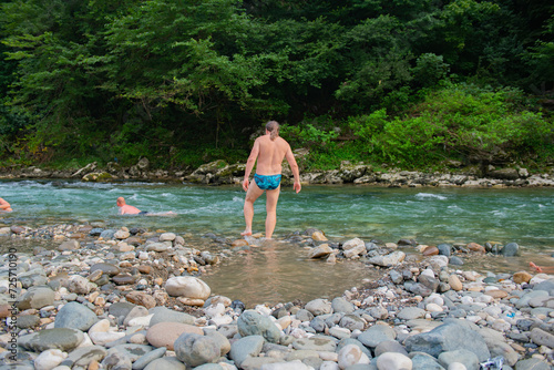 Man with Long Hair Swimming in Scenic Mountain River with Trees and Stones in Background
