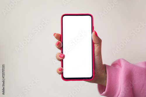 Close-up, no face, female hand in pink manicure sweatshirt pink nails holding smartphone in pink case with white blank screen for mockup or copy space on pastel gray background. Use of Applications