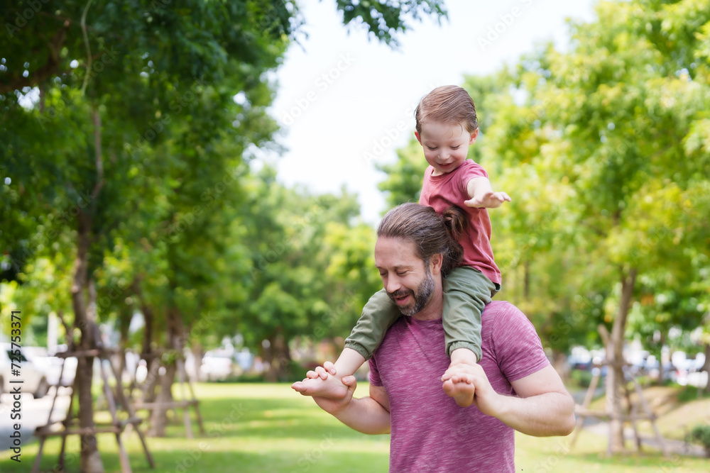 Fototapeta premium Handsome Caucasian man, taking my little son play in garden on nice day, son was placed on neck, little boy placed arms on father's head, father held onto cute son's legs for safety.