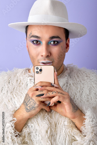 Stylish womanlike man LGBTQ chatting with friend on phone posing at camera, isolated on purple studio background. caucasian european guy with make-up in white fluffy coat. beauty, fashion