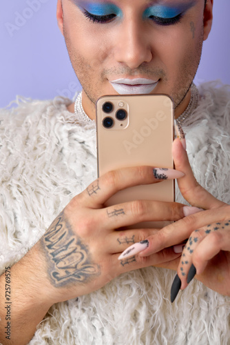 Confident womanlike man LGBTQ chatting with someone on phone posing at camera, smiling, isolated on purple studio background. caucasian european guy with make-up in white fluffy coat. close-up