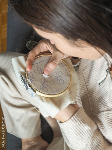Detail left-handed woman embroiders at home in pajamas. Close-up view of an unrecognizable left-handed person sitting cross-legged on a dark gray sofa. She is embroidering small white flowers