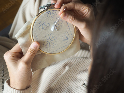 Detail view of two hands embroidering in round bamboo frame. An unrecognizable person is embroidering some white flowers on a beige fabric. The woman is in lounge clothes, comfortable, on her sofa.