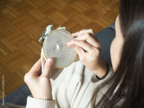 Detail view of a woman embroidering in the tranquility of her house. A calm person sitting on the couch at home embroidering with a needle on beige fabric using white thread following a flower pattern