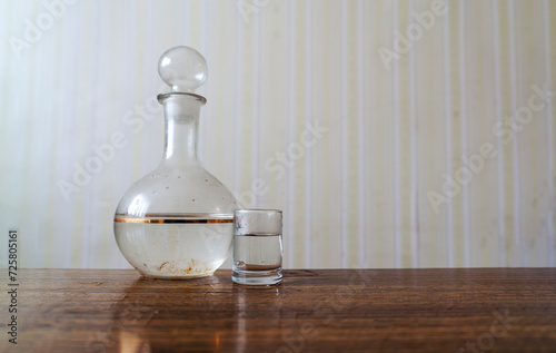 Glass dirty retro decanter with vodka and one shot located on wooden table in apartment with old vintage wallpaper on background