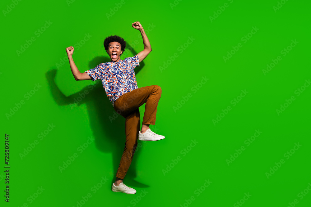 Fulll length photo of lucky cheerful guy wear print shirt yelling yes rising fists empty space isolated green color background Fulll length photo of lucky cheerful guy wear print shirt yelling yes rising fists empty space isolated green color background