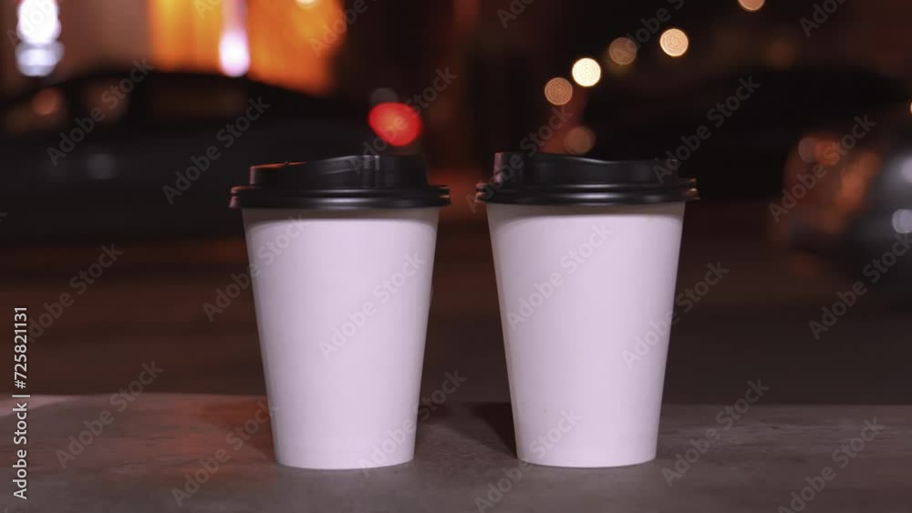 Two white paper disposable cups with black lids on blurred background of evening city lights. Hot Takeaway Coffee Outdoors. Street food, urban lifestyle concept. Copy space, space for text. Two white paper disposable cups with black lids on blurred background of evening city lights. Hot Takeaway Coffee Outdoors. Street food, urban lifestyle concept. Copy space, space for text.