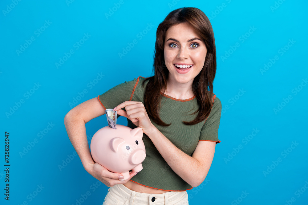 Photo of lovely cheerful girl toothy smile hold put dollar bill money bank pig isolated on blue color background Photo of lovely cheerful girl toothy smile hold put dollar bill money bank pig isolated on blue color background