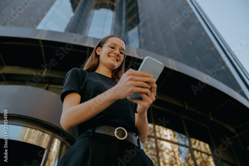 Bottom view of business woman in eyeglases use mobile phone while standing on building background photo