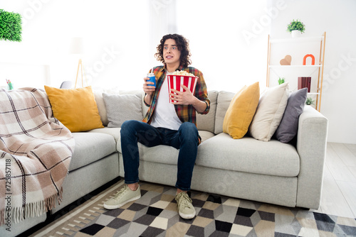 Full length photo of excited funny guy wear plaid shirt eating pop corn can coke watching interesting movie indoors room home house