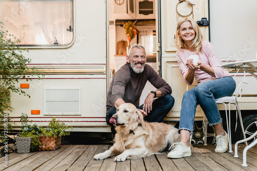 Portrait of senior joyful smiling european couple with their labrador resting in the doorway of their caravan home. Traveling with domestic animals golden retriever