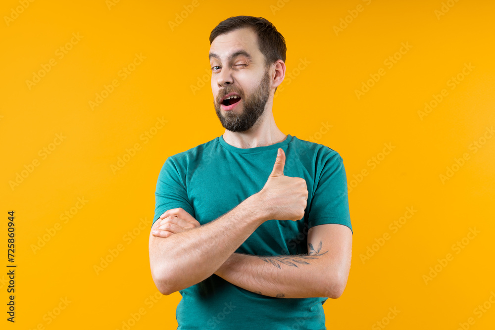Studio portrait of confident young man posing over bright colored yellow background winking and holding thumb up - obrazy, fototapety, plakaty 