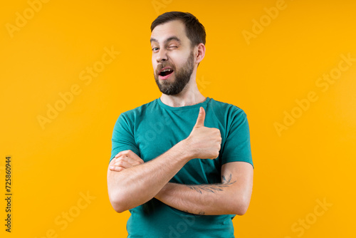 Studio portrait of confident young man posing over bright colored yellow background winking and holding thumb up photo