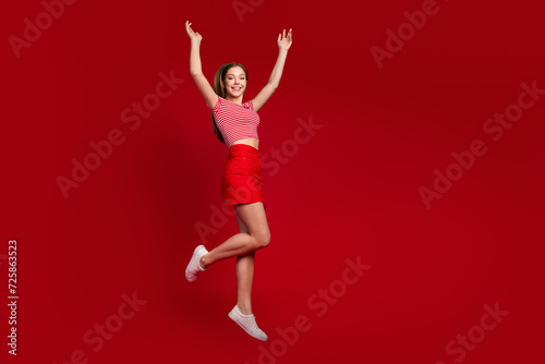 Full length photo of funny excited lady dressed striped top mini skirt jumping high rising hands arms empty space isolated red color background