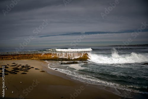 2023-12-31 THE CHILDRENS POOL IN LA JOLLA CALIFORNIA WITH WAVES CRASHING ON THE BRICK RETAINING WALL WITH SEA LIONS ON THE SAND AND A CLOUDY SKY photo