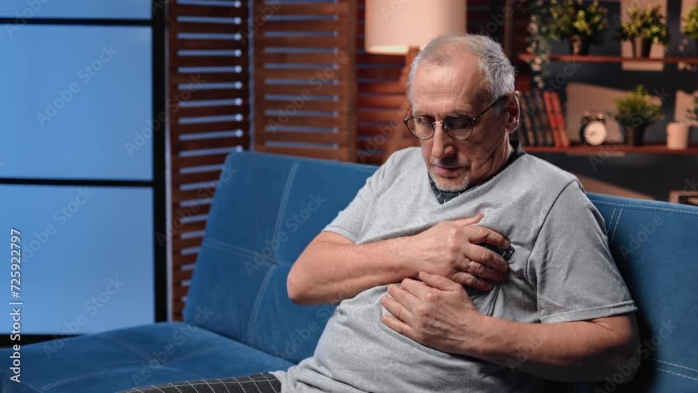 Portrait of elderly man with heartache sitting on couch in home, holding left part of chest by hand Portrait of elderly man with heartache sitting on couch in home, holding left part of chest by hand