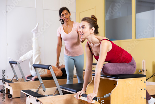 Friendly Hispanic woman professional pilates instructor helping young girl exercising on combo chair in fitness studio