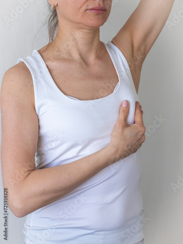 Shot of the woman in the white top against the white wall, performing self examination of the breasts, looking for abnormalities. Cancer awareness