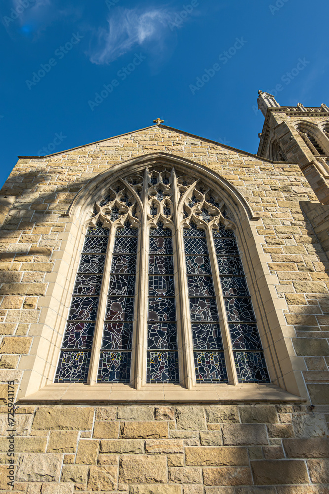 Naklejka premium Santa Barbara, CA, USA - November 30, 2023: Trinity Episcopal Church, closeup of large stained glass window set in sculpted stone frame on State Street. Part of tower. Blue cloudscape