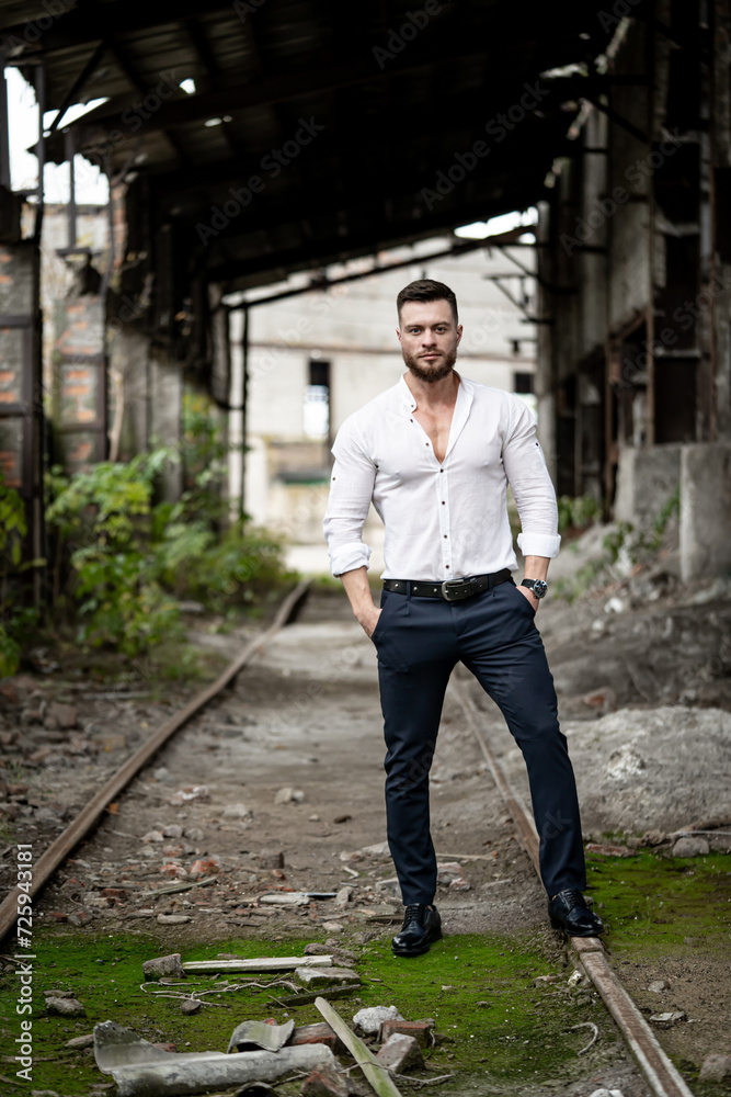 Handsome man model of fashion with beard and stylish hair dressed in white shirt and black pants in abandoned area near the railway