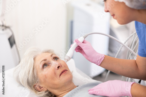 Pleased aged woman having vacuum cleaning of her face by means of apparatus for aesthetic procedures used by specialist photo