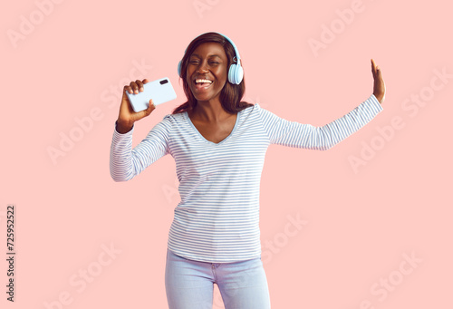 Happy cheerful joyful positive young African woman on pastel studio background uses mobile phone and noise cancellation headphones head set, listens to music, has good time, enjoys her favorite tracks