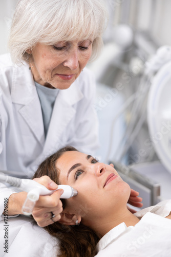 Careful old woman doctor conducting vacuum cleaning of young woman patient s face in aesthetic clinic