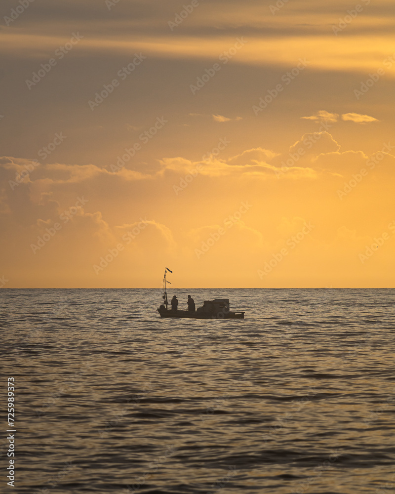 
A boat sailing on the sea with clouds and the horizon in the background. Silhouette of a fishing boat sailing alone in the vastness of the sea at sunrise.